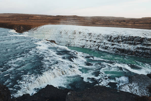 The iconic Gullfoss waterfall in Iceland flows through a partially frozen landscape, capturing the transition between autumn and winter. The surrounding terrain is tinged with warm orange hues, while snow and ice cling to the rocks and cliffs around the cascading waters. Under a pastel-hued sunset, the sky glows with soft purples and oranges, adding a serene, almost ethereal quality to this powerful natural wonder.