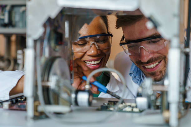 A mid-adult Caucasian male and a diverse female scientist smile as they adjust robotics equipment in a high-tech laboratory, their expertise evident in their precise movements.
