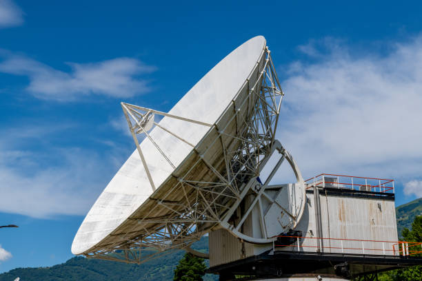 Gravedona, Lombardei - Italy - 06-24-2024: Large satellite dish with a white surface pointing into the blue sky, surrounded by forested hills in the background.
