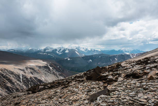 istock Dramatic top view from stone pass above alpine valley to large mountain range with snow-capped peaks on horizon in gray cloudy sky. Sunlit rocky steep slope and snowy tops far away under rainy clouds. 2183770626