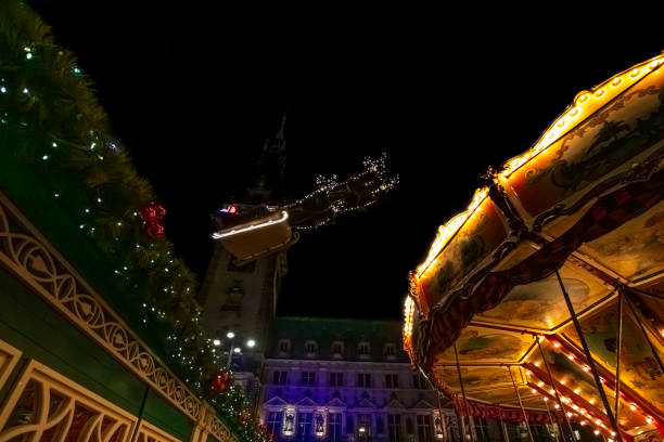 Hamburg, Germany – December 21, 2023: A merry-go-round and Christmas decorations, seen in the foreground, are all lit up at night, while Santa’s sleigh flies above the Hamburger Weihnachtsmarkt Rathausmarkt, a traditional Christmas market in the northern German city of Hamburg.