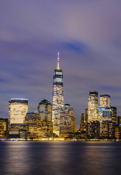Manhattan’s iconic skyline at twilight glows with illuminated skyscrapers like the Empire State and One World Trade Center, reflected in the Hudson River. The soft blue sky and sparkling city lights capture the city's vibrant energy and architectural grandeur.
