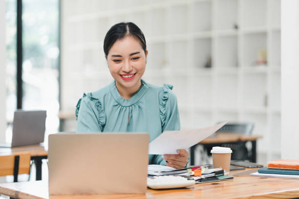 istock woman is smiling while working on laptop in modern office environment, holding documents in her hand. atmosphere is bright and productive, reflecting positive work experience. 2183858244