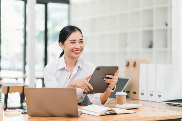 istock young woman is sitting at wooden desk, smiling while using tablet. She is dressed in white shirt and is surrounded by laptop, notebook, and coffee cup, creating productive and cheerful workspace 2183858464