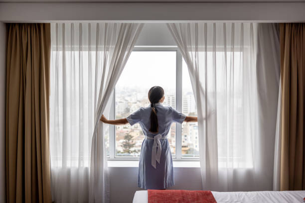 istock Maid working at a hotel and opening the drapes in a room while looking through the window 2183861991