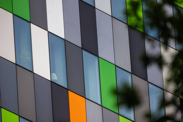 istock The photograph shows a modern building facade with a grid of rectangular panels in shades of green, white, orange, and grey. Some windows reveal interior elements, with a leafy tree branch partially in view. 2183869274