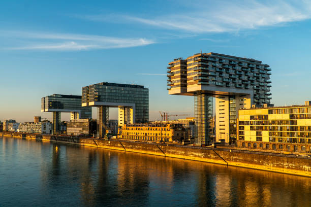 A modern cityscape featuring three unique crane-shaped buildings along the river, with a bridge in front carrying a train. The buildings reflect the warm light of the setting sun.