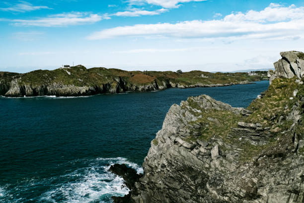 istock Waves gently lap against the rugged cliffs while a lighthouse stands proudly in the distance, surrounded by lush greenery under a bright blue sky. 2183876091