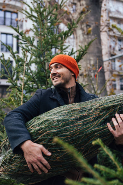 istock Man Joyfully Carrying Christmas Tree in Urban Setting 2183898465