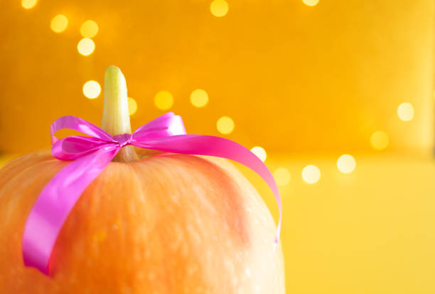 A pumpkin with a pink ribbon tied around it. The pumpkin is sitting on a yellow background. The ribbon is tied in a bow, giving the pumpkin a festive and playful appearance
