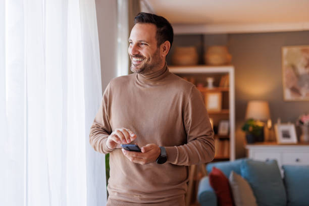 istock Happy mid adult man with mobile phone looking through window thoughtfully while standing in living room 2183988561