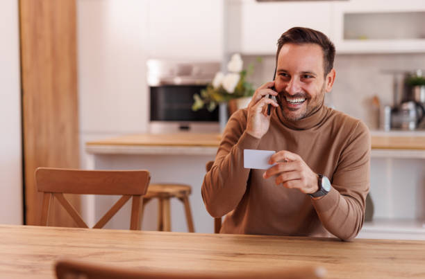 istock Smiling mid adult man with credit card talking over mobile phone while sitting at dining table 2183988611