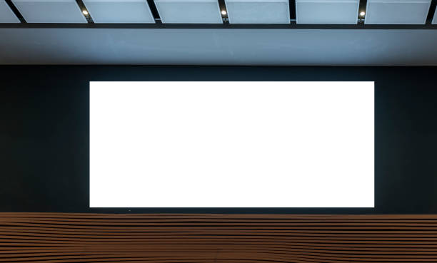 istock Blank white screen mounted on black wall in modern interior space with wood panel accents at bottom. Ceiling lights and white ceiling panels add minimalist feel to setting 2183991494