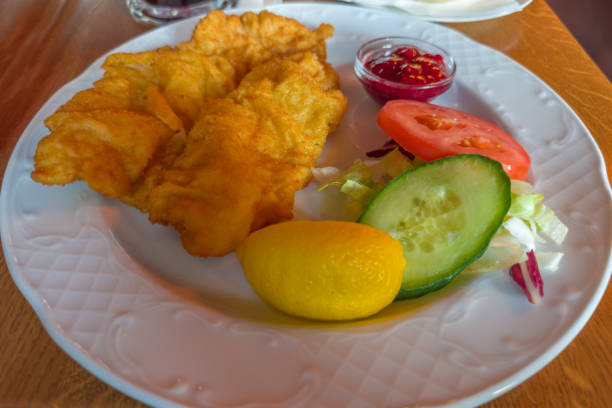 Serving of roasted flat thin boneless breaded pork chop, known as Schnitzel Viennese style with lemon and fresh vegetables on the white dish on a wooden table