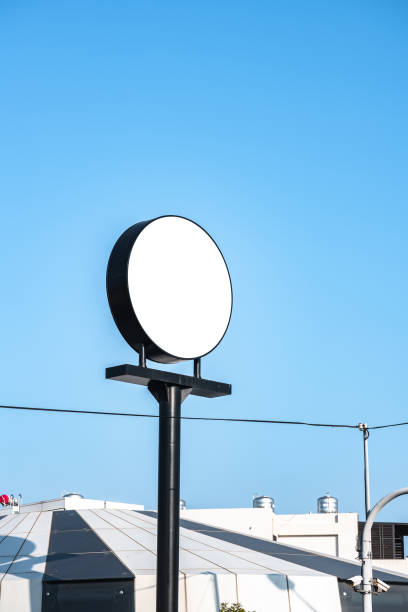 Circular blank sign elevated against a clear blue sky, providing ample space for customization. Industrial rooftops and wires form the background, offering an urban setting. Ideal for conveying messages or branding concepts with a minimalist touch.