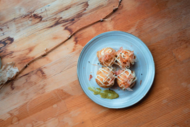 Tempting Takoyaki Delight Deliciously plated takoyaki served on a round ceramic dish, showcasing perfectly cooked octopus balls garnished with bonito flakes and drizzled with creamy sauce. The rustic wooden table adds warmth, complementing the appetizing dish, while wasabi provides a touch of color and spice on the side. cooking takoyaki is a ball shaped japanese snack stock pictures, royalty-free photos & images