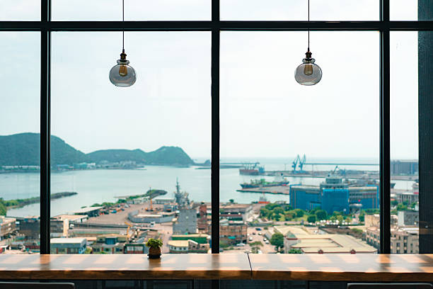 Overlooking a bustling harbor city. Large glass windows frame the picturesque scene, featuring industrial cranes and distant hills under a clear sky. Warm lighting from elegant pendant lamps creates a cozy ambiance, contrasting with the vibrant urban landscape outside.