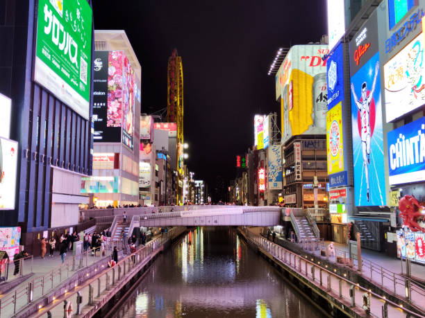People walking on Dotonbori canal waterfront by the central Ebisu Bridge with many illuminated signboards. Dotonbori is a famous commercial and nightlife area in downtown Osaka, Japan.