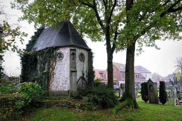 A small, ivy-covered chapel with a pointed roof stands in a cemetery surrounded by trees and gravestones. The chapel is made of brick and has circular windows.