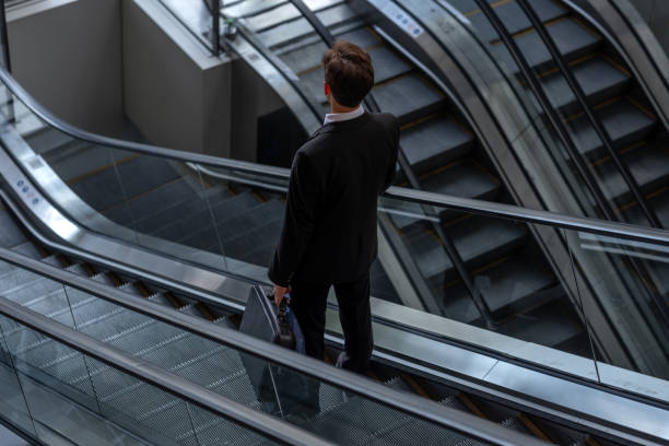 istock Young businessman standing on escalators with phone and briefcase in hand 2184014818