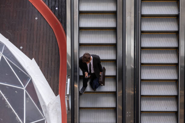 istock Young businessman standing on escalators with phone and briefcase in hand 2184014836