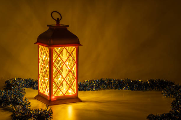 A cozy holiday setup featuring a glowing lantern casting warm light, surrounded by a decorative blue tinsel garland. The warm tones and gentle glow create a festive and inviting atmosphere, ideal for Christmas decor.