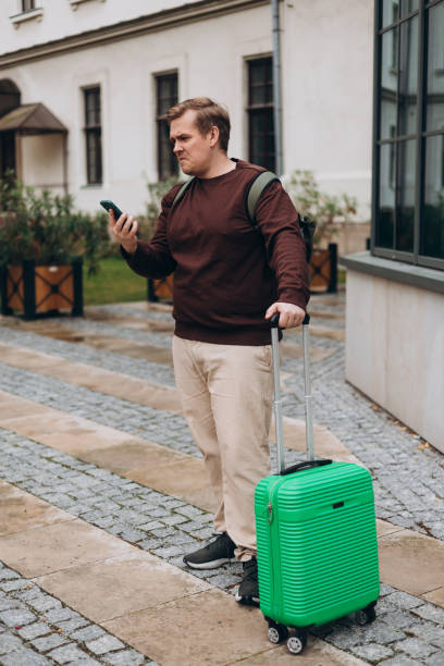 istock Upset sad, skeptical man holding smartphone gadget for communicate outdoors. Negative people emotion, Urban lifestyle concept. Angry 30s male with green suitcase reading bad news, full body 2184026798