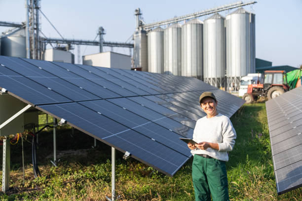 istock Female farmer wearing work uniform holds digital tablet in her hands stands next to solar panels. Modern agricultural farm using solar panels. Agricultural silos in the background. 2184049964