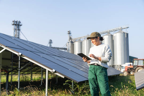 istock Female farmer wearing work uniform holds digital tablet in her hands stands next to solar panels. Modern agricultural farm using solar panels. Agricultural silos in the background. 2184050423