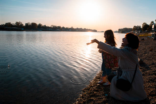 Mother and little daughter throwing pebbles in the lake, enjoying the sunset on the beach in Autumn. Copy space