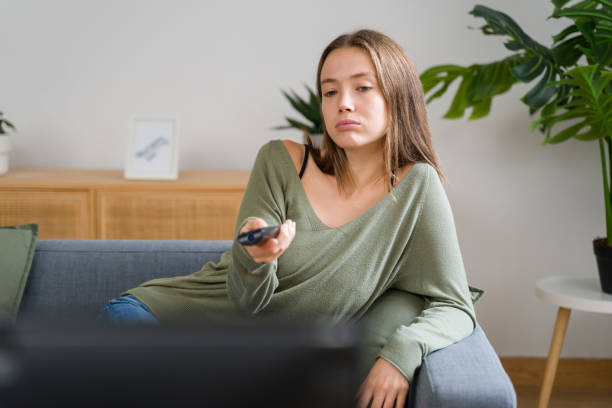 istock Bored young woman changing channels while watching television on sofa at home 2184057096