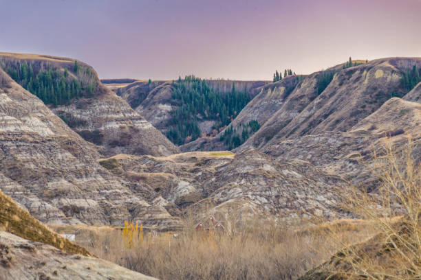 A scenic view of rugged, layered hills and valleys at dusk, with sparse trees scattered on slopes and a few evergreen clusters at the top. A red barn nestles in the valley below, adding a touch of rural charm to the vast, earthy landscape