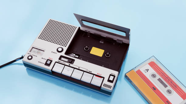 Vintage cassette tape recorder and an audio cassette tape against a light blue background. The recorder has buttons for play, record, and other functions, with a compartment open for loading a cassette. The setup evokes a retro or nostalgic aesthetic, reminiscent of the 1980s or early 1990s.