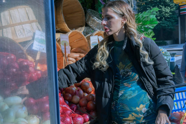 A side photograph of a pregnant woman outdoors selecting groceries