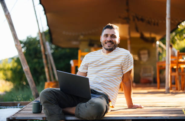 istock Man Relaxing Outdoors with Laptop at Rustic Cabin Retreat 2184069330