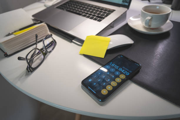 A laptop, a cell phone, a cup of coffee, and a stack of papers are on a table. The laptop is open and the cell phone is placed next to it