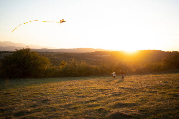 A helping hand. Caring mother teaches little girl to fly a kite in autumn nature at sunset. Child development, learning, freedom, joy, & connecting with nature.