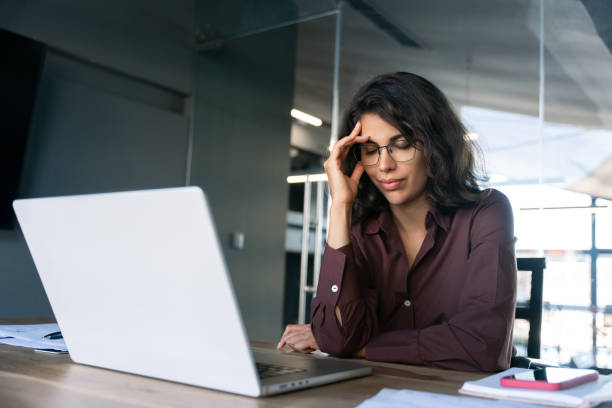 istock Focused young businesswoman looking at laptop pc computer screen having headache, migraine. Latin business woman holding hand near temples, feeling stressed and tired sitting at workplace in office 2184105567