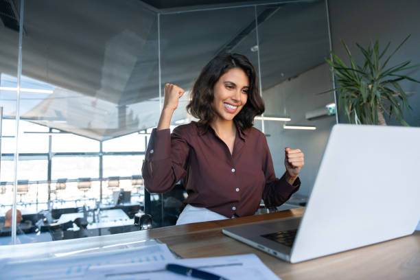Happy excited young businesswoman looking at laptop pc computer screen doing yes triumph win gesture. Smiling latin hispanic business woman successful winner celebrating winning in office. Copy space