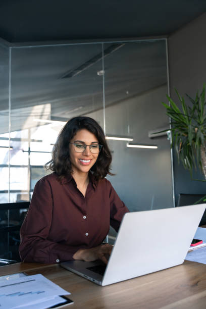 istock Smiling young professional it specialist latin hispanic business lady working on laptop pc sitting in modern office space. Middle eastern indian woman using computer technology app for work. Vertical 2184105573