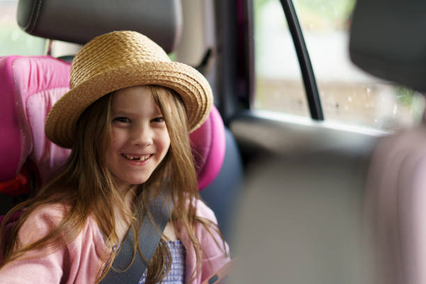 istock Little cute smiling girl without a front tooth in a sarafan and a straw hat sits fastened with a belt in a car. The concept of children's travel and recreation, health and dentistry 2184105696