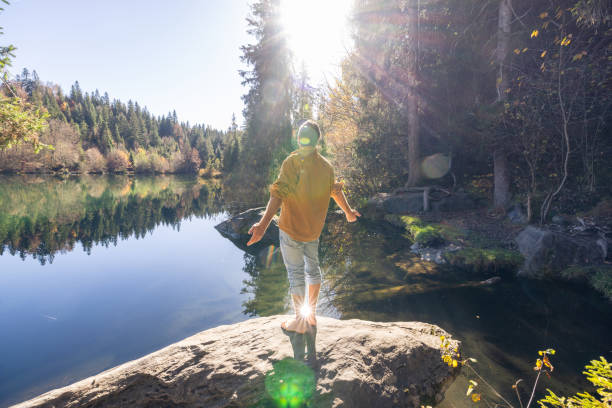 A man stands barefoot with arms wide open, facing a calm lake and dense forest, embracing the peaceful atmosphere of the outdoors.
