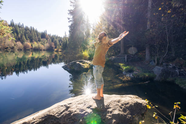 A man stands barefoot with arms wide open, facing a calm lake and dense forest, embracing the peaceful atmosphere of the outdoors.