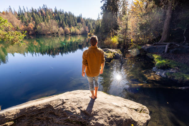A man stands barefoot on a rock overlooking a serene lake surrounded by lush forest, embracing nature’s beauty and calmness.