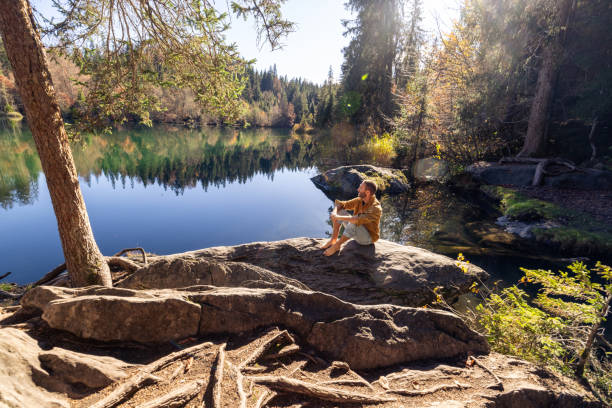 Smiling man sits barefoot on a rock by a peaceful forest lake, enjoying the calm and natural beauty of the autumn landscape.