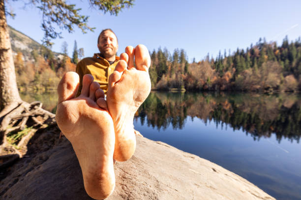 A close-up of bare feet in focus with a man relaxing by a lake surrounded by autumn trees. A unique perspective captures the essence of relaxation and connection to nature.