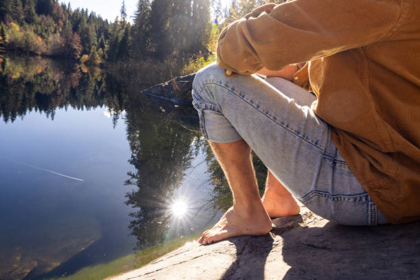 Close-up view of a man sitting barefoot by a calm lake with sunlight reflecting on the water. The image captures a peaceful moment in nature, focusing on his  feet.