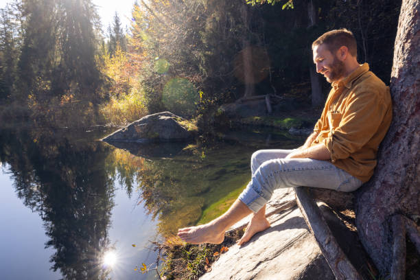 A man leans against a tree, eyes closed in peaceful contemplation beside a serene lake surrounded by nature. Sunlight filters through the trees, adding a warm glow.