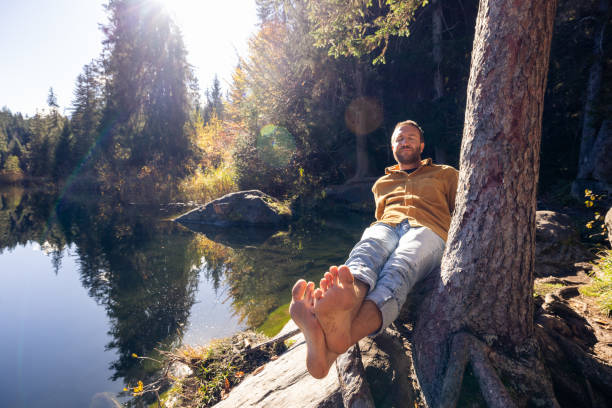 A man sits barefoot by a calm lake, leaning against a tree with a content smile. Sunlight filters through the trees, highlighting the peaceful, natural setting.