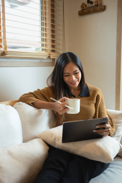 istock Charming young woman holding cup of hot beverage using digital tablet on sofa 2184113881